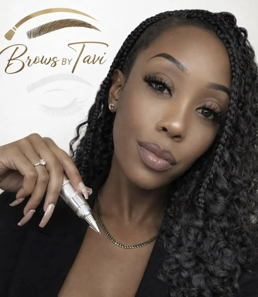 Tavi, semi-permanent makeup artist, holding a PMU pen in front of the Brows by Tavi neon logo